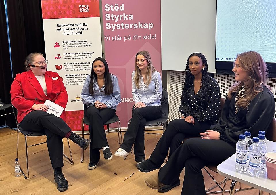 Fyra unga tjejer samtalar med en kvinna i röd kavaj. Alla sitter ned framför två rollups. Den ena med texten: Stöd. "Styrka. Systerskap."