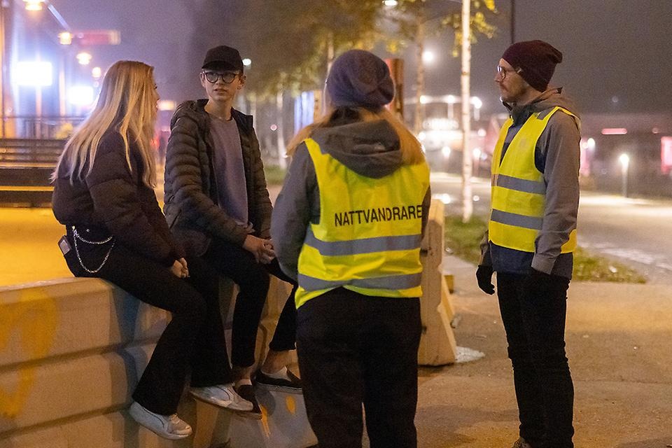 Två vuxna med nattvandringsvästar talar med ungdomar utomhus på kvällen.