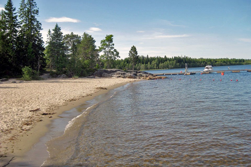 Små vågor som slår mot strandkanten, en båtbrygga i bakgrunden.