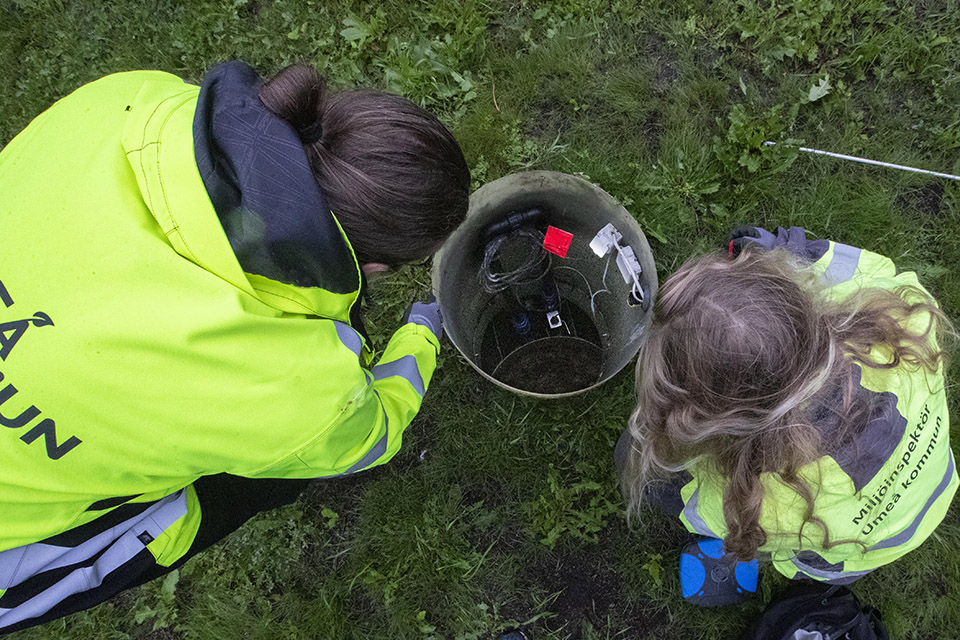 Två personer med varselkläder sitter böjda över en avloppsanläggning och tittar ner i ett rör.