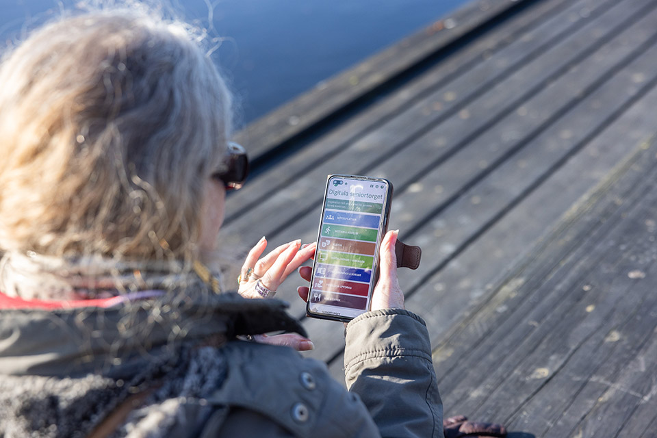 En äldre kvinna som är porträtterad bakifrån, hon håller i en mobiltelefon med Digitala seniortorget öppet.