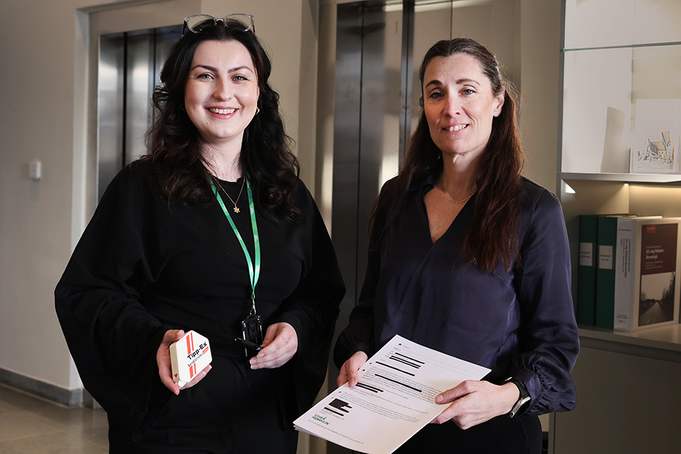 Cornelia och Malin står bredvid varandra i stashusets reception. De håller upp ett utskrivet maskerat dokument och verktyg som används för maskering på det traditionella sättet.