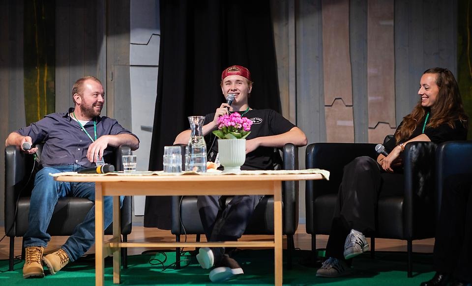 Tre glada personer i fåtöljer. Den unga mannen i mitten håller i en mikrofon.